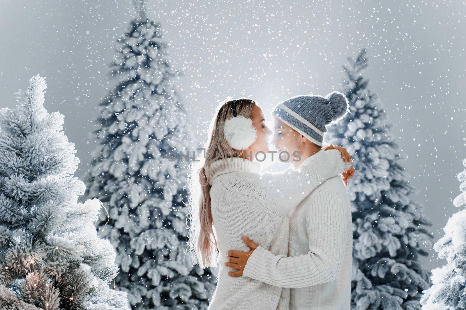 Falling snow and kisses. Happy young couple close-up hugs and kiss near christmas trees at the eve of new year celebration in winter day. Smiley man and woman weared white pullovers love each other