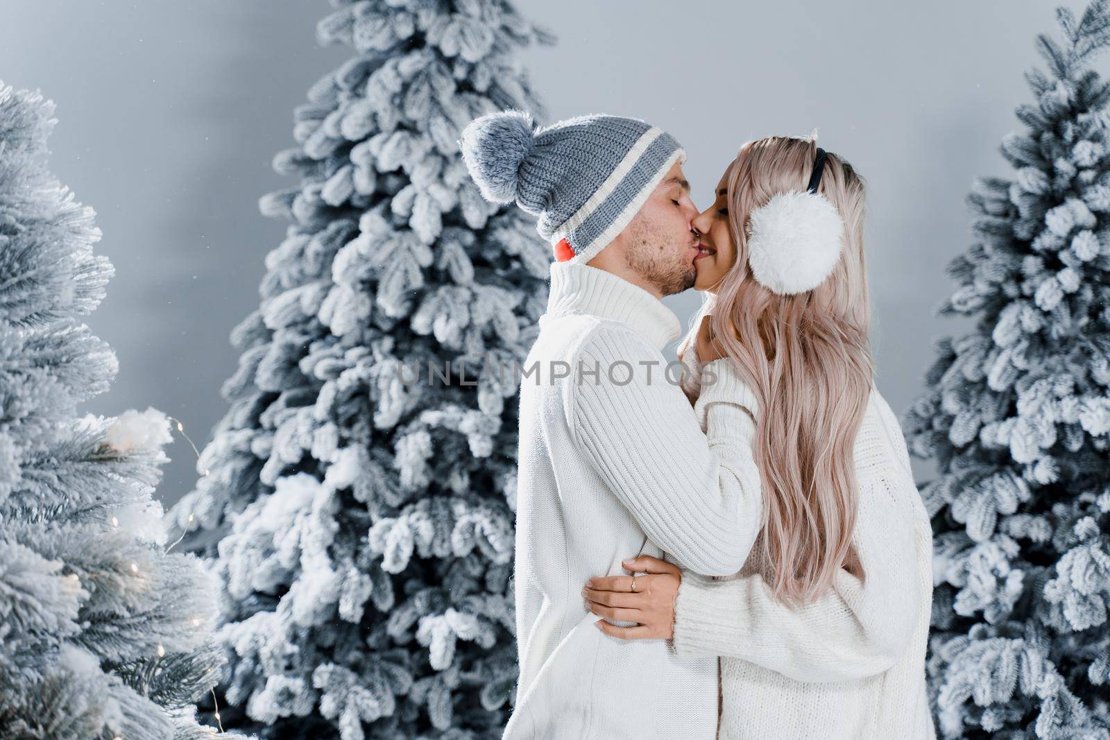 Couple kissing near christmass trees. Winter holidays. Love story of young couple weared white pullovers. Happy man and young woman hug each other.