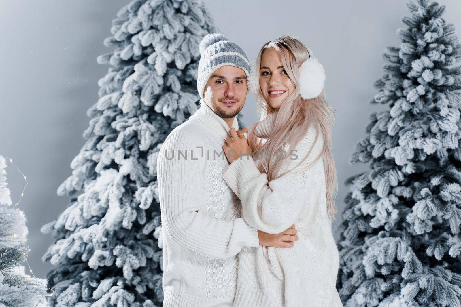 Winter love story with couple weared fur headphones, hats, white sweaters. Happy young couple hugs and kiss near christmas trees at the eve of new year celebration in winter day.