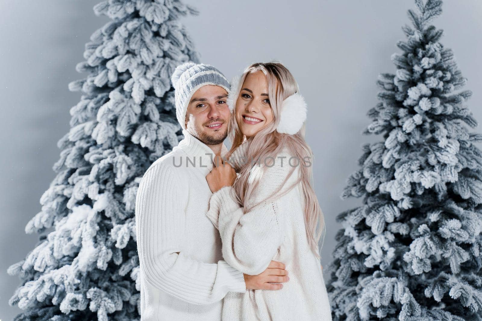 Winter love story with couple weared fur headphones, hats, white sweaters. Happy young couple hugs and kiss near christmas trees at the eve of new year celebration in winter day by Rabizo