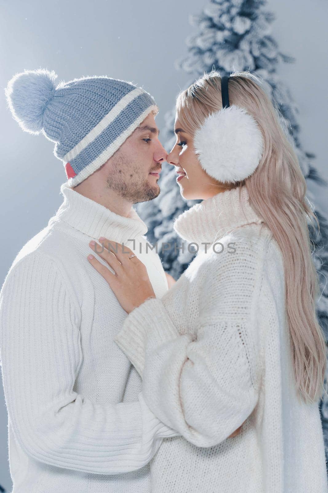 Winter love story with couple weared fur headphones, hats, white sweaters. Happy young couple hugs and kiss near christmas trees at the eve of new year celebration in winter day.