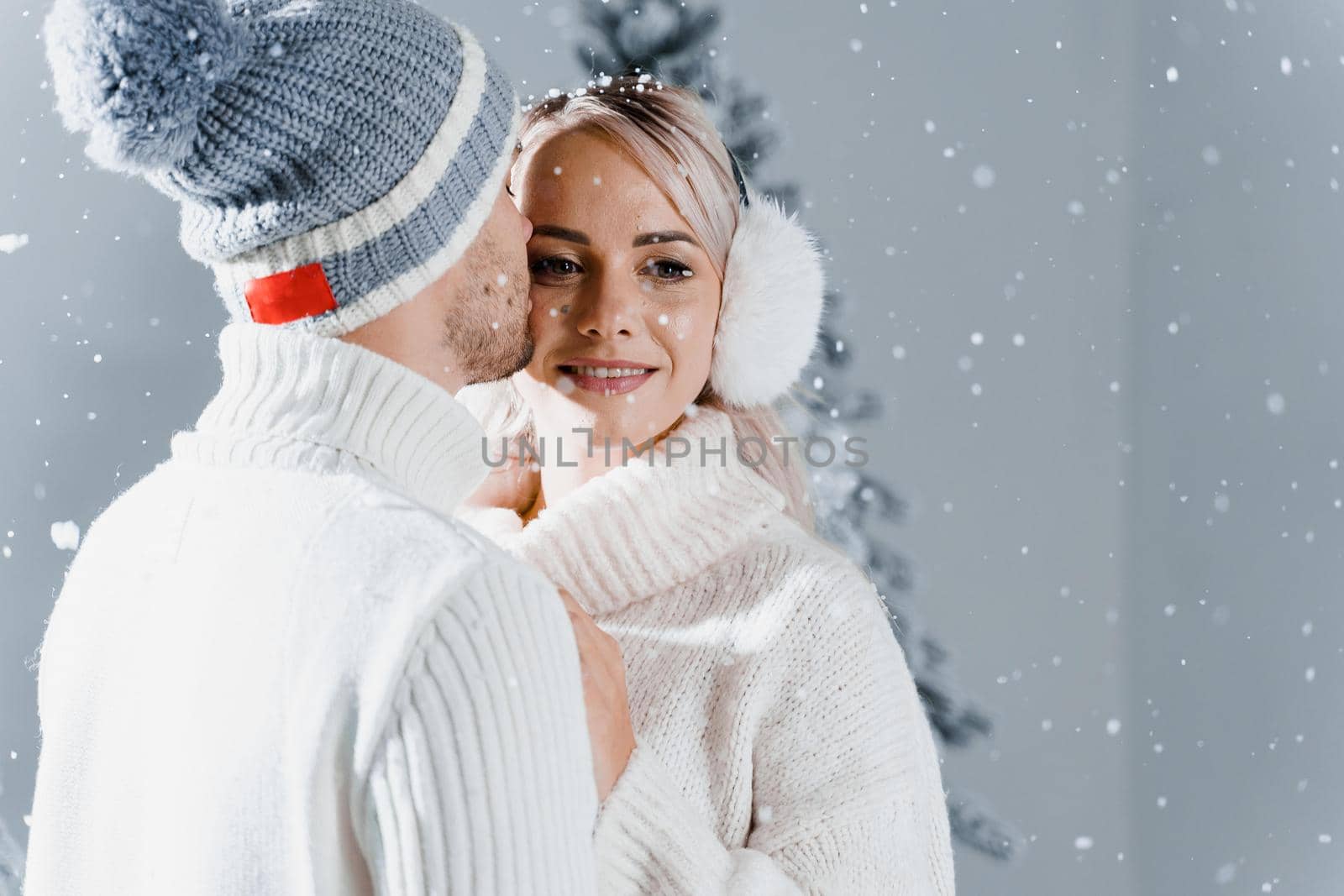 Happy young couple hugs and smiles and snow falls near christmas trees at the eve of new year celebration in winter day. Smiley man and woman weared white pullovers love each other