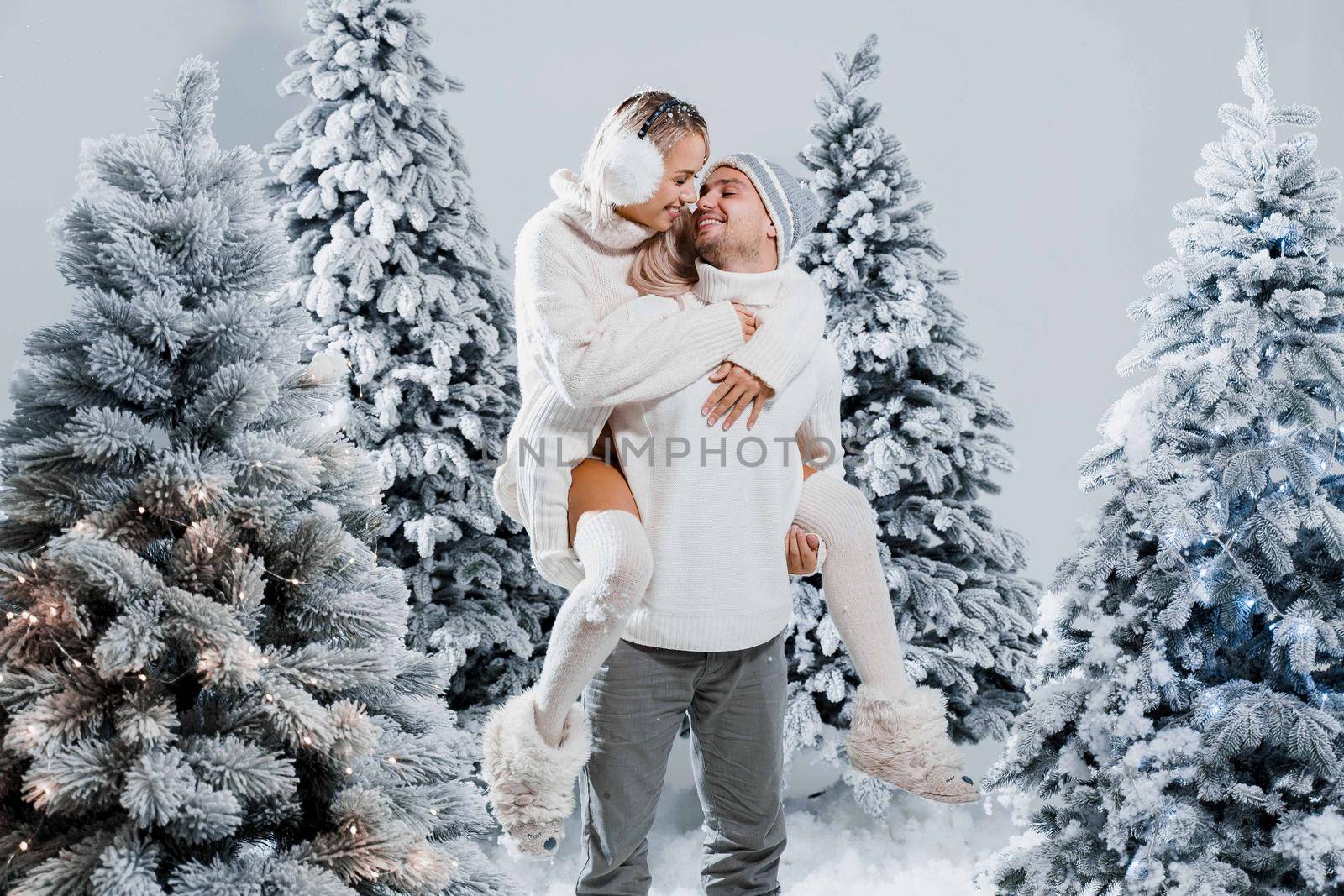 Couple kiss and hug. Man holds girl near christmas trees in winter day. New year celebration. People weared wearing fur headphones, hats, white sweaters by Rabizo