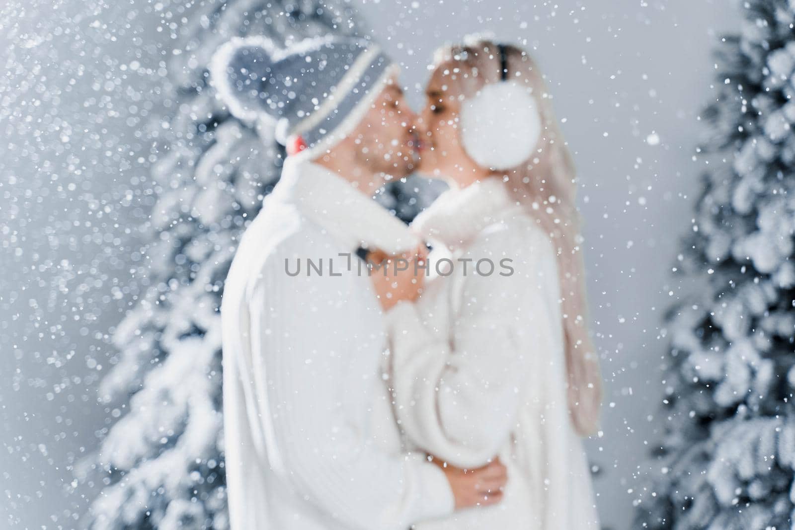 Happy young couple hugs and smiles and snow falls near christmas trees at the eve of new year celebration in winter day. Smiley man and woman weared white pullovers love each other