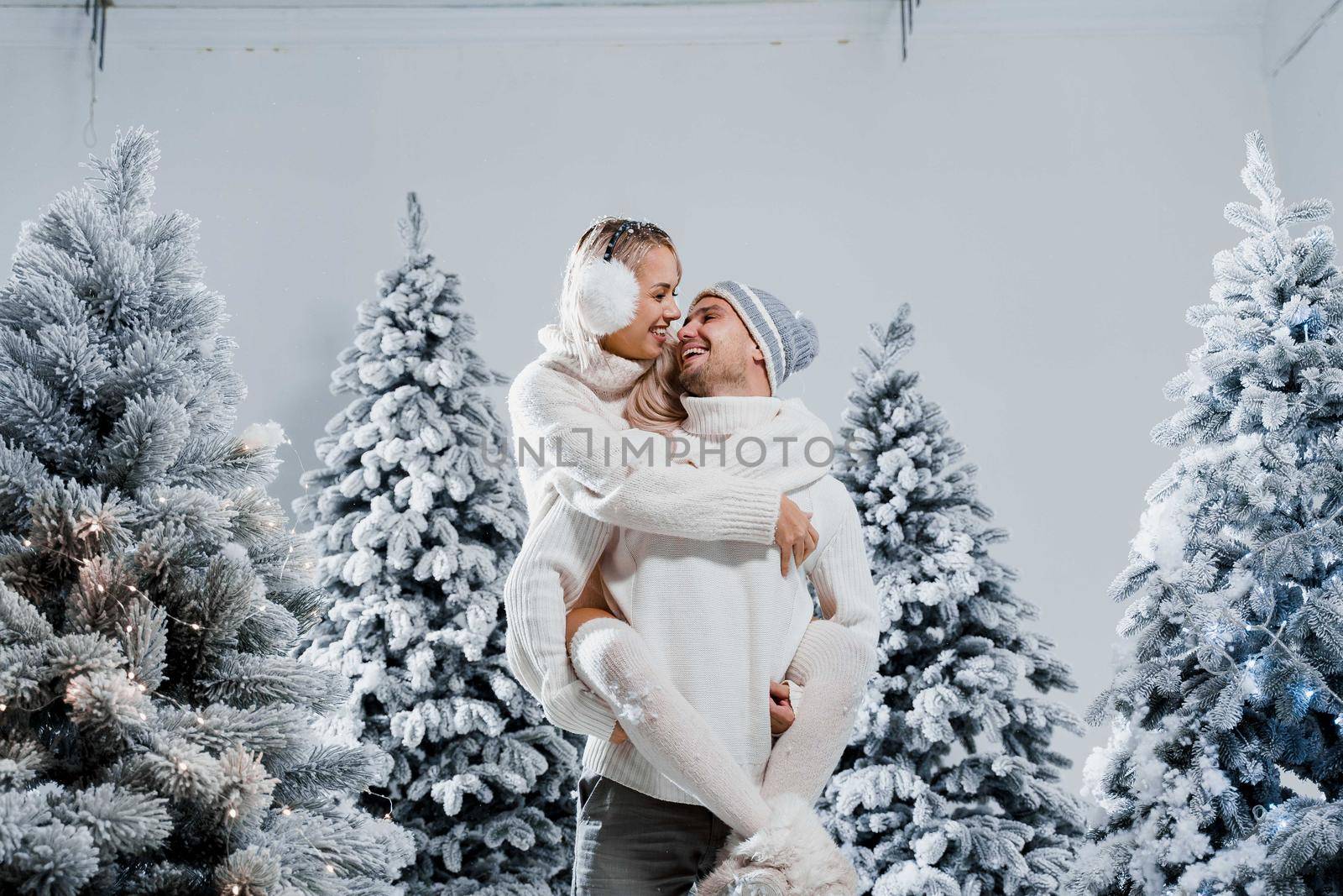 Couple kiss and hug. Man holds girl near christmas trees in winter day. New year celebration. People weared wearing fur headphones, hats, white sweaters by Rabizo