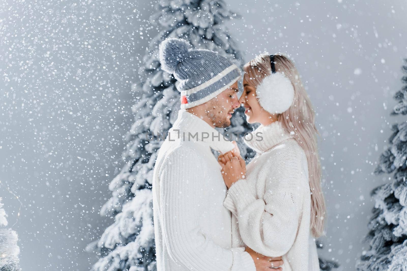 Happy young couple hugs and smiles and snow falls near christmas trees at the eve of new year celebration in winter day. Smiley man and woman weared white pullovers love each other