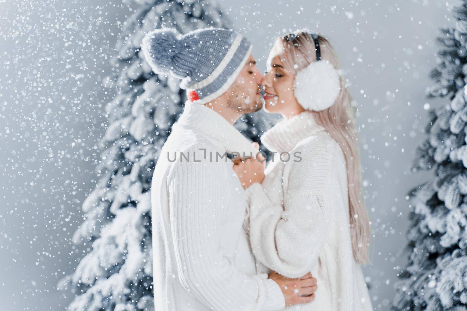 Couple kissing while snow falls near christmass trees. Winter holidays. Love story of young couple weared white pullovers. Happy man and young woman hug each other.