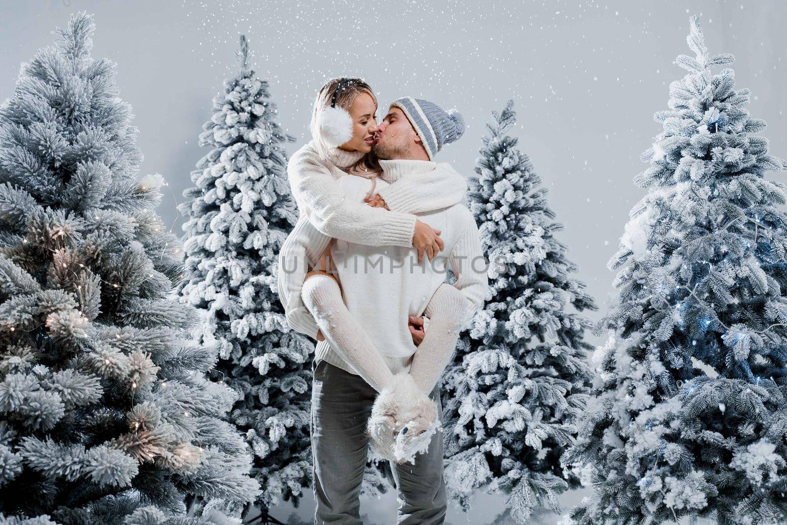 Couple kiss and hug. Man holds girl near christmas trees in winter day. New year celebration. People weared wearing fur headphones, hats, white sweaters.