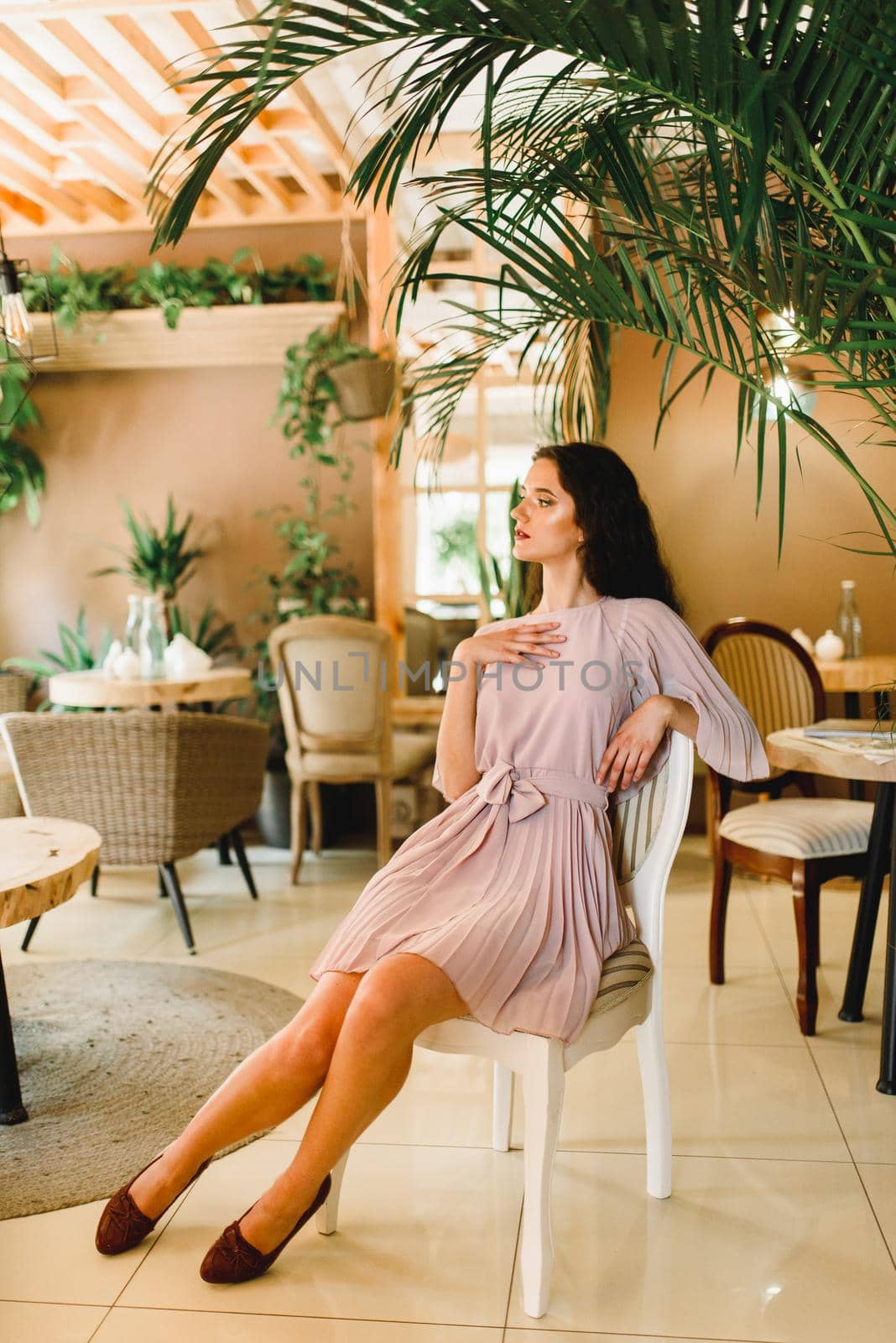 Young woman in soft pink dress is sitting on the chair and looking to the left side. Beauty girl in cafe with big green plants