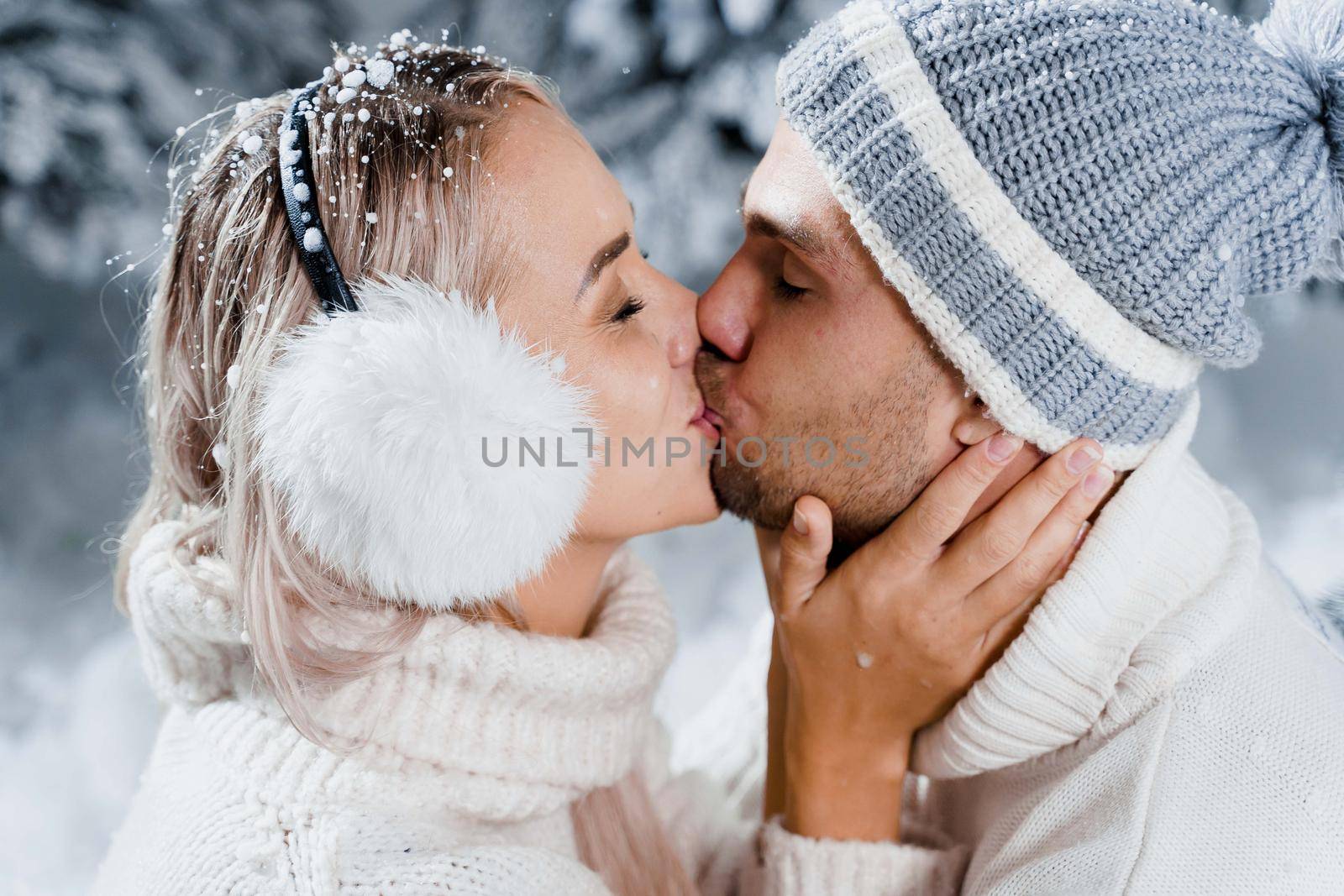 New year love story. Couple kisses and hugs. People weared wearing fur headphones, hats, white sweaters. Happy young couple hugs and kiss near christmas trees in winter day.