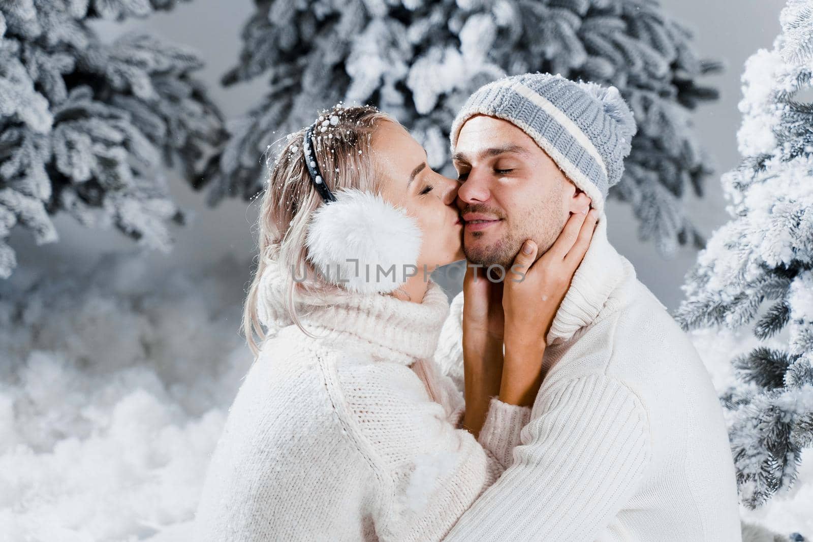 Winter love story with couple weared fur headphones, hats, white sweaters. Happy young couple hugs and kiss near christmas trees at the eve of new year celebration in winter day by Rabizo