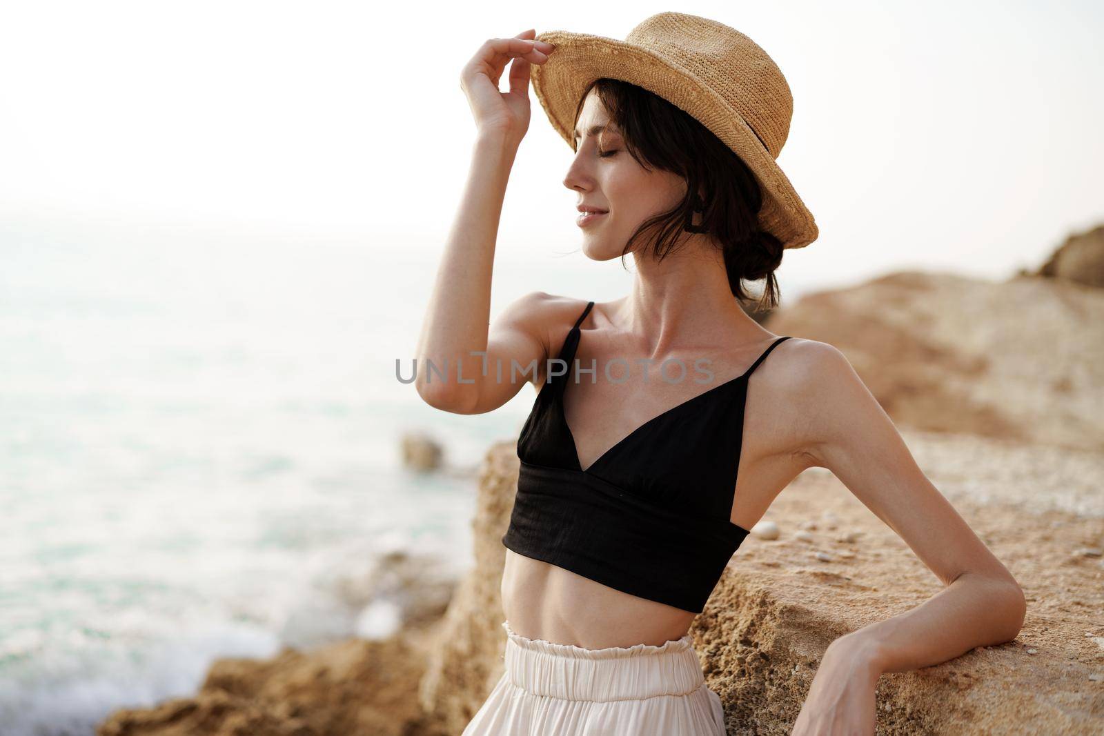 Portrait of woman in black bralette and white trousers leaning on rock at beach by Fabrikasimf