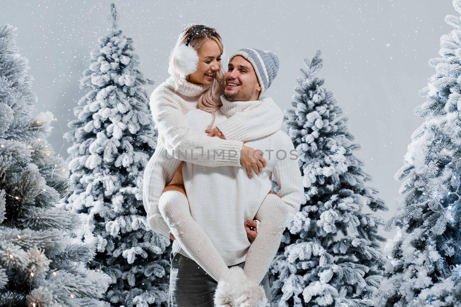 Couple kiss and hug. Man holds girl near christmas trees in winter day. New year celebration. People weared wearing fur headphones, hats, white sweaters.