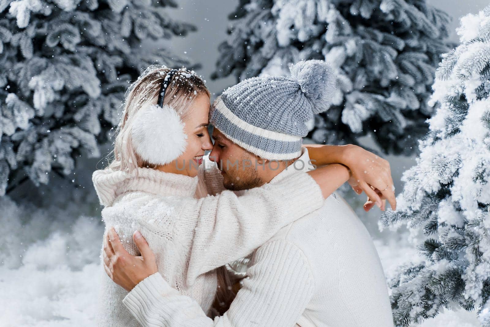 Winter love story with couple weared fur headphones, hats, white sweaters. Happy young couple hugs and kiss near christmas trees at the eve of new year celebration in winter day.