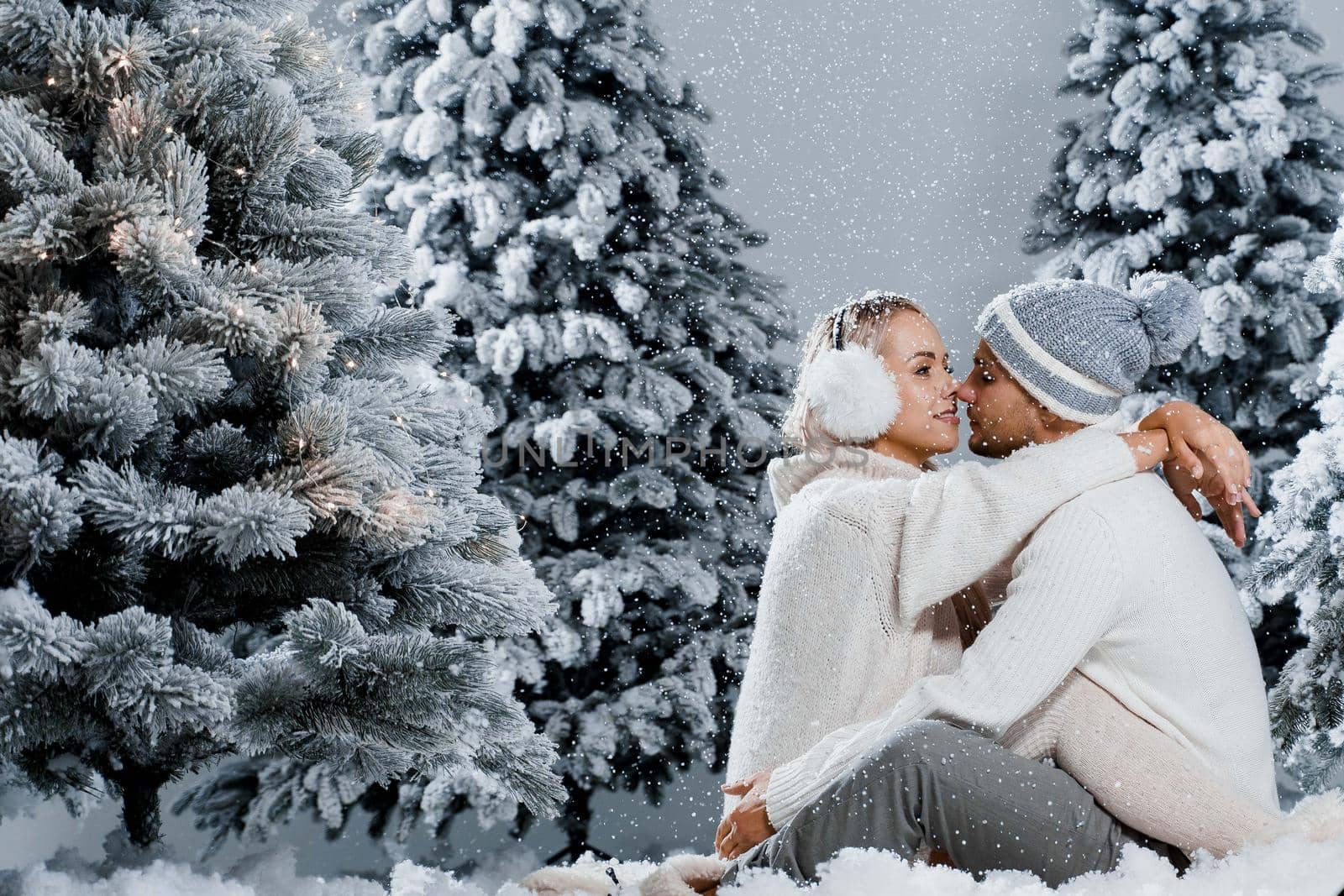 Couple seat on the snow and hug, kiss, and have fun each other. Winter love story before new year celebration. Waiting for christmas gift. Happy couple weared fur headphones, hats, white sweaters by Rabizo