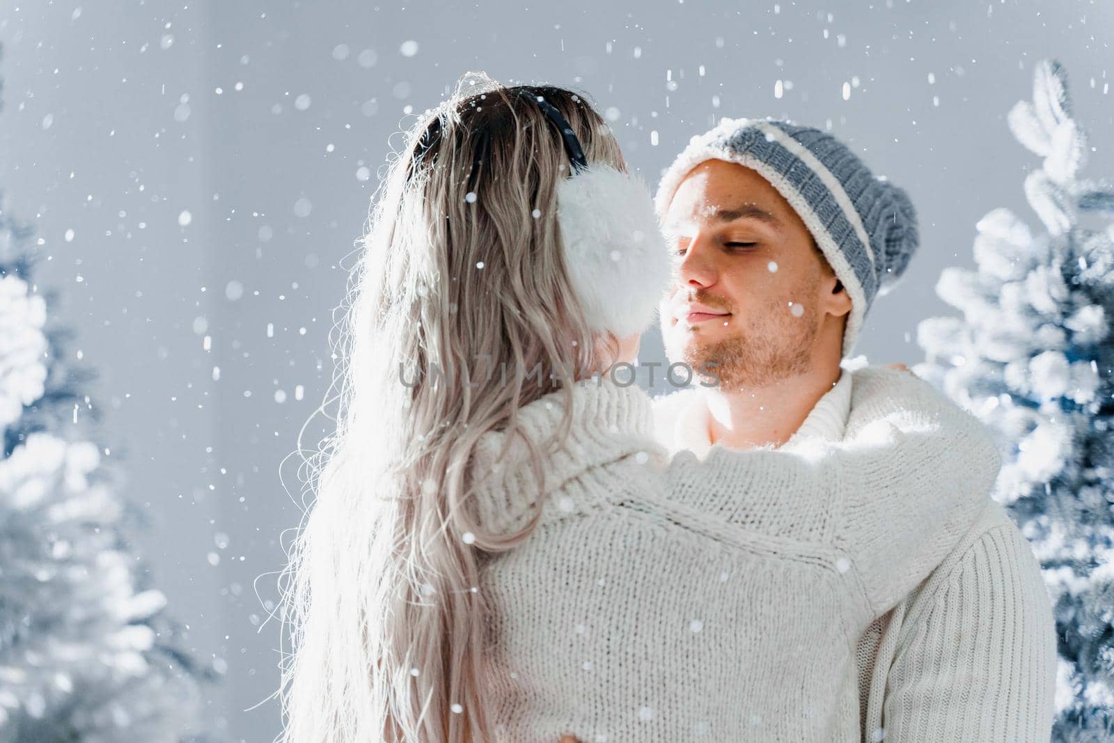 Happy young couple hugs and smiles and snow falls near christmas trees at the eve of new year celebration in winter day. Smiley man and woman weared white pullovers love each other