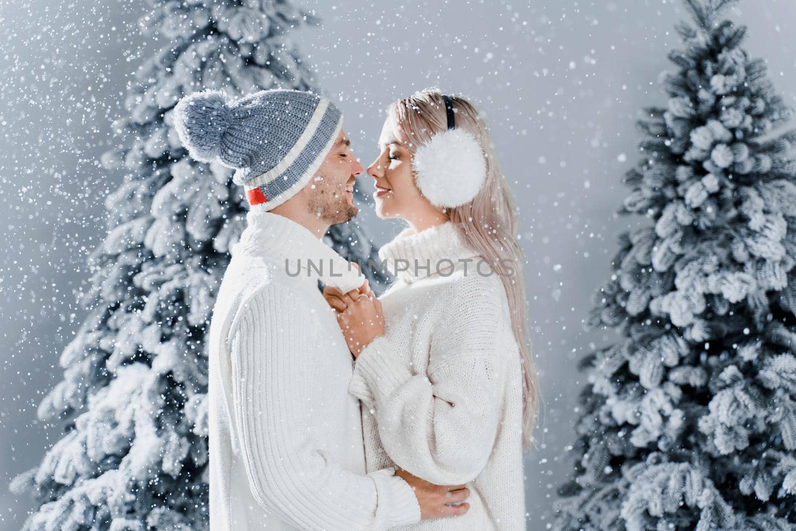 Happy young couple hugs and smiles and snow falls near christmas trees at the eve of new year celebration in winter day. Smiley man and woman weared white pullovers love each other, by Rabizo