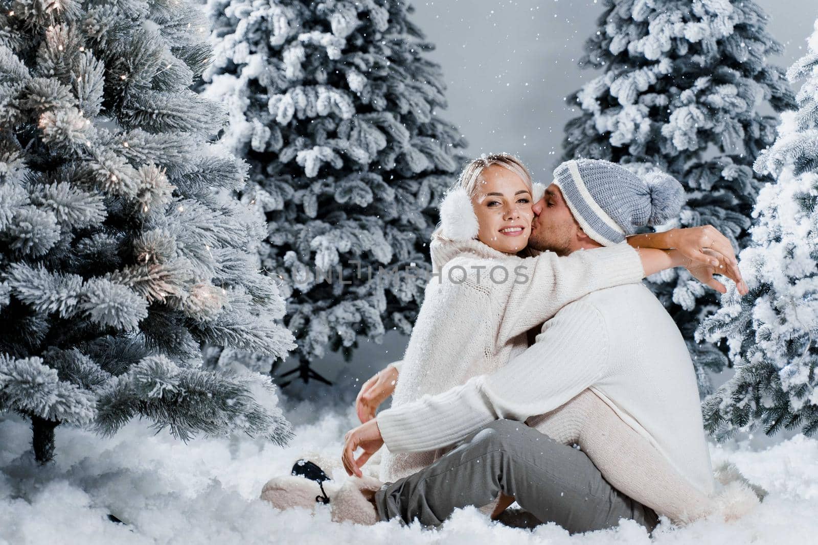 Couple seat on the snow and hug, kiss, and have fun each other. Winter love story before new year celebration. Waiting for christmas gift. Happy couple weared fur headphones, hats, white sweaters.