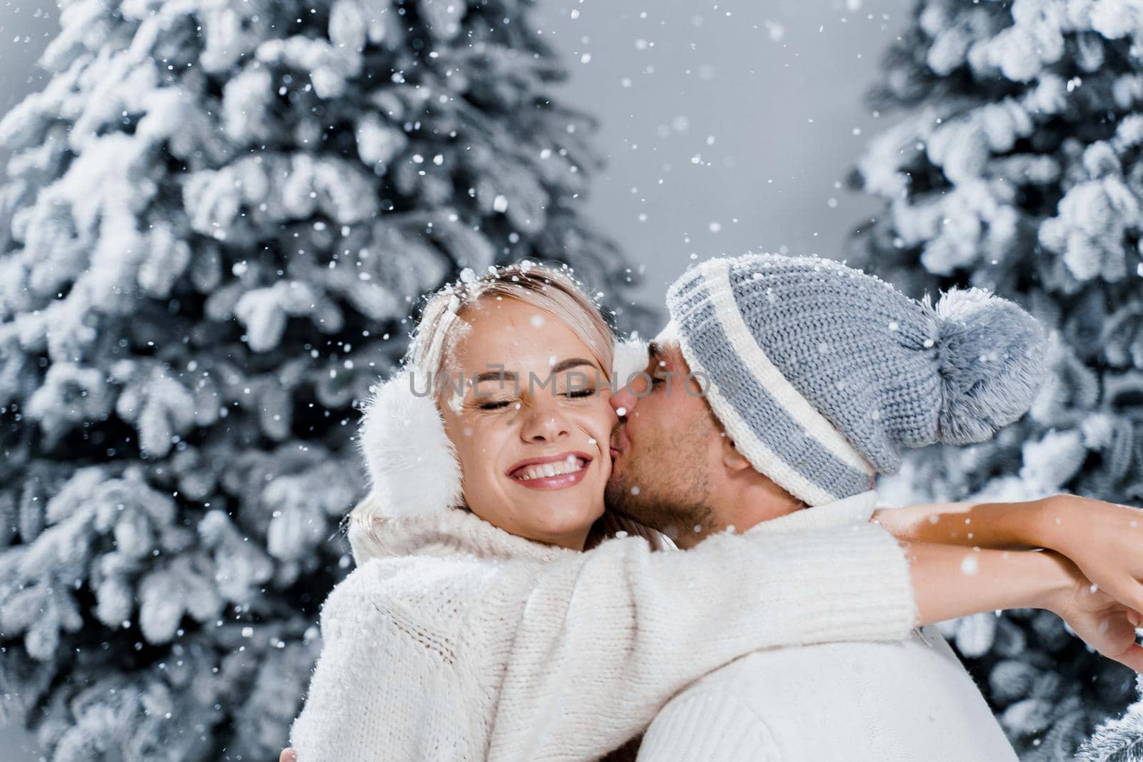 Couple seat on the snow and hug, kiss, and have fun each other. Winter love story before new year celebration. Waiting for christmas gift. Happy couple weared fur headphones, hats, white sweaters by Rabizo