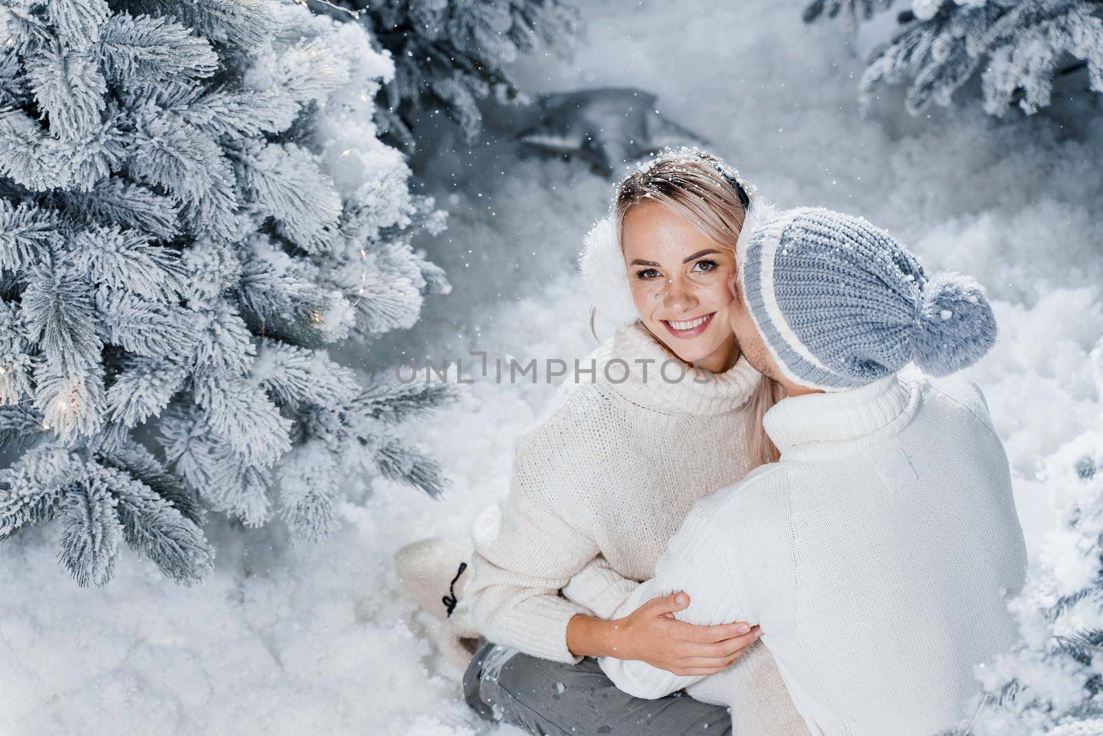 Man kiss and hug his woman and snow falls. New year love story. People weared wearing fur headphones, hats, white sweaters by Rabizo