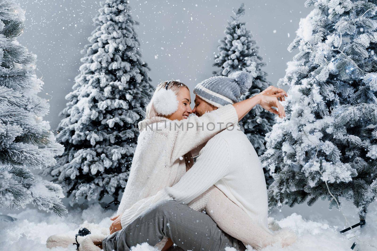 Falling snow and kisses. Happy young couple close-up hugs and kiss near christmas trees at the eve of new year celebration in winter day. Smiley man and woman weared white pullovers love each other, by Rabizo