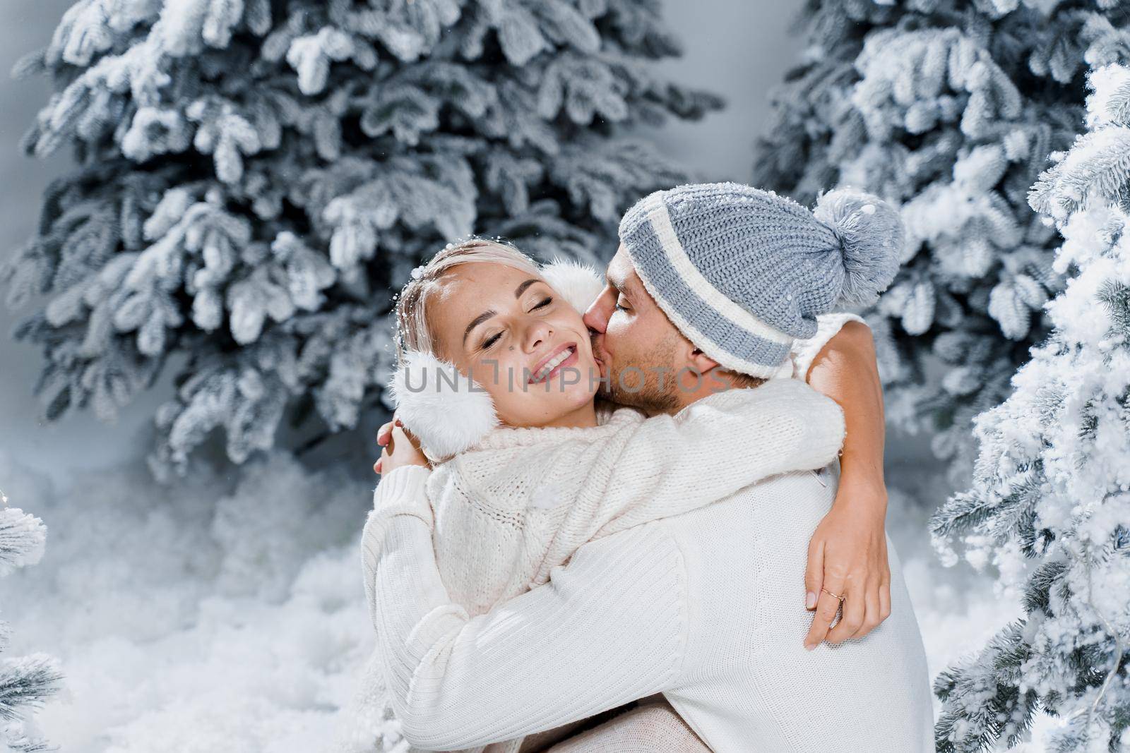 Handsome man is kissing his lovely girl in cheek. Winter holidays. Love story of young couple weared white pullovers. Happy man and young woman hug each other. by Rabizo