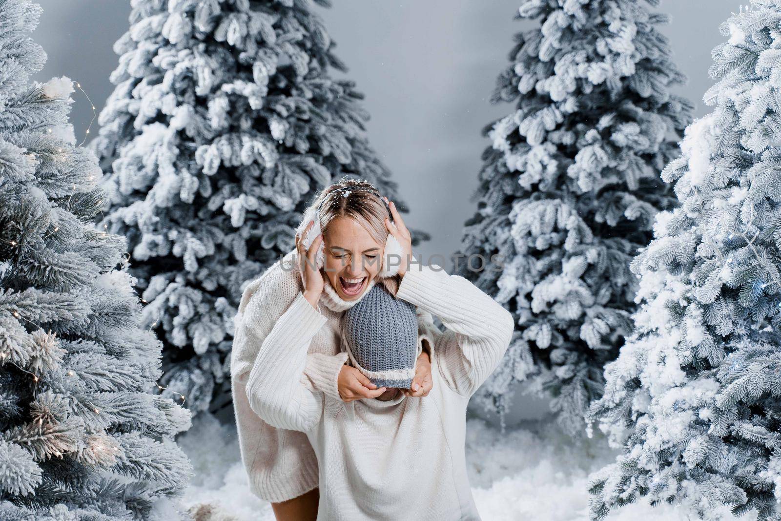 Falling snow and kisses. Happy young couple close-up hugs and kiss near christmas trees at the eve of new year celebration in winter day. Smiley man and woman weared white pullovers love each other