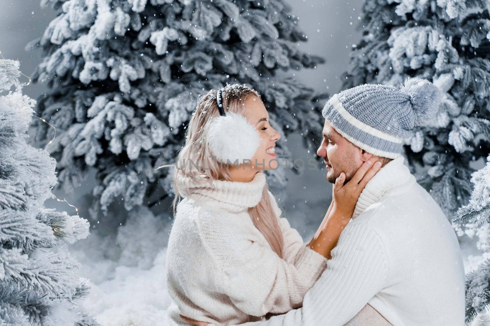 Couple seat on the snow and hug, kiss, and have fun each other. Winter love story before new year celebration. Waiting for christmas gift. Happy couple weared fur headphones, hats, white sweaters.