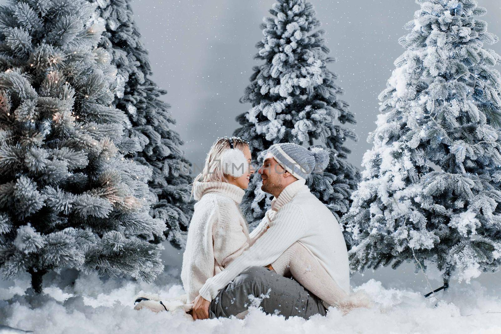 Man kiss and hug his woman and snow falls. New year love story. People weared wearing fur headphones, hats, white sweaters.