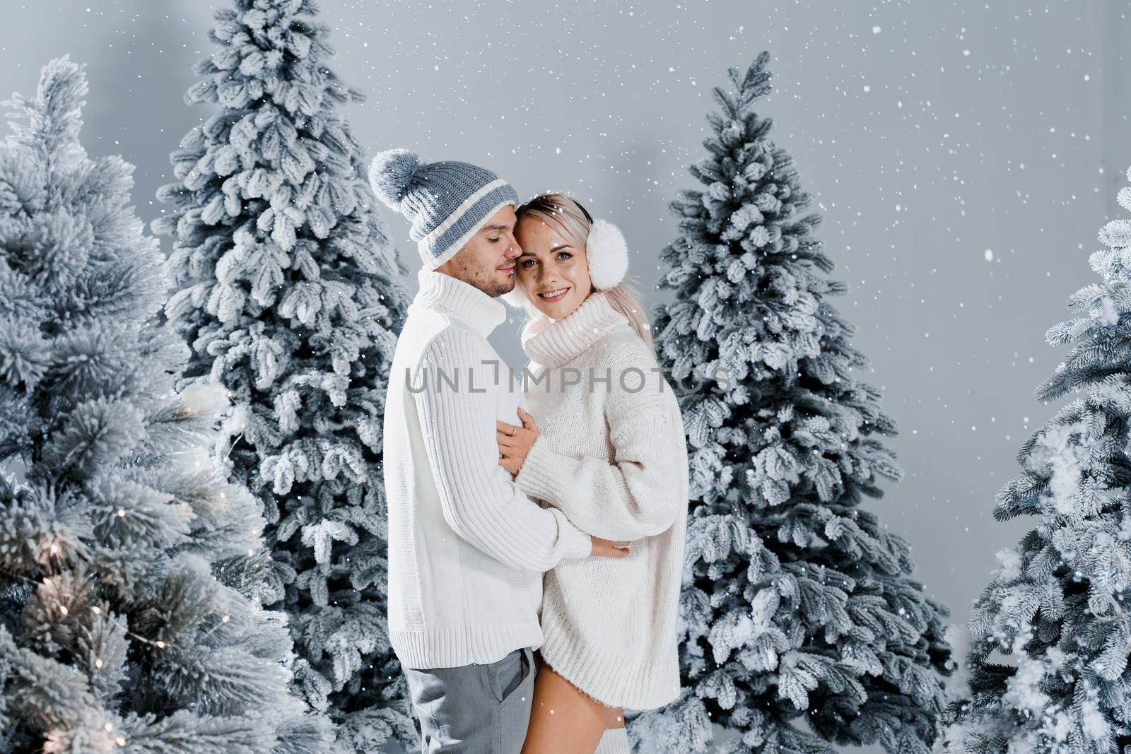 Couple hugging near christmass trees. Winter holidays. Love story of young couple weared white pullovers. Happy man and young woman love each other.