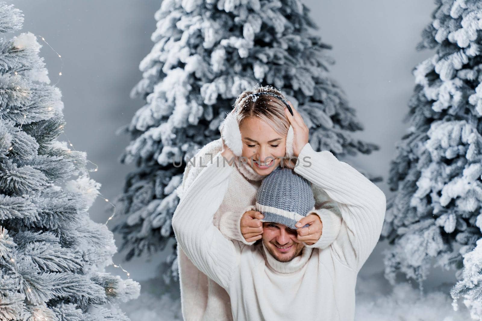 Winter love story with couple weared fur headphones, hats, white sweaters. Happy young couple hugs and kiss near christmas trees at the eve of new year celebration in winter day by Rabizo