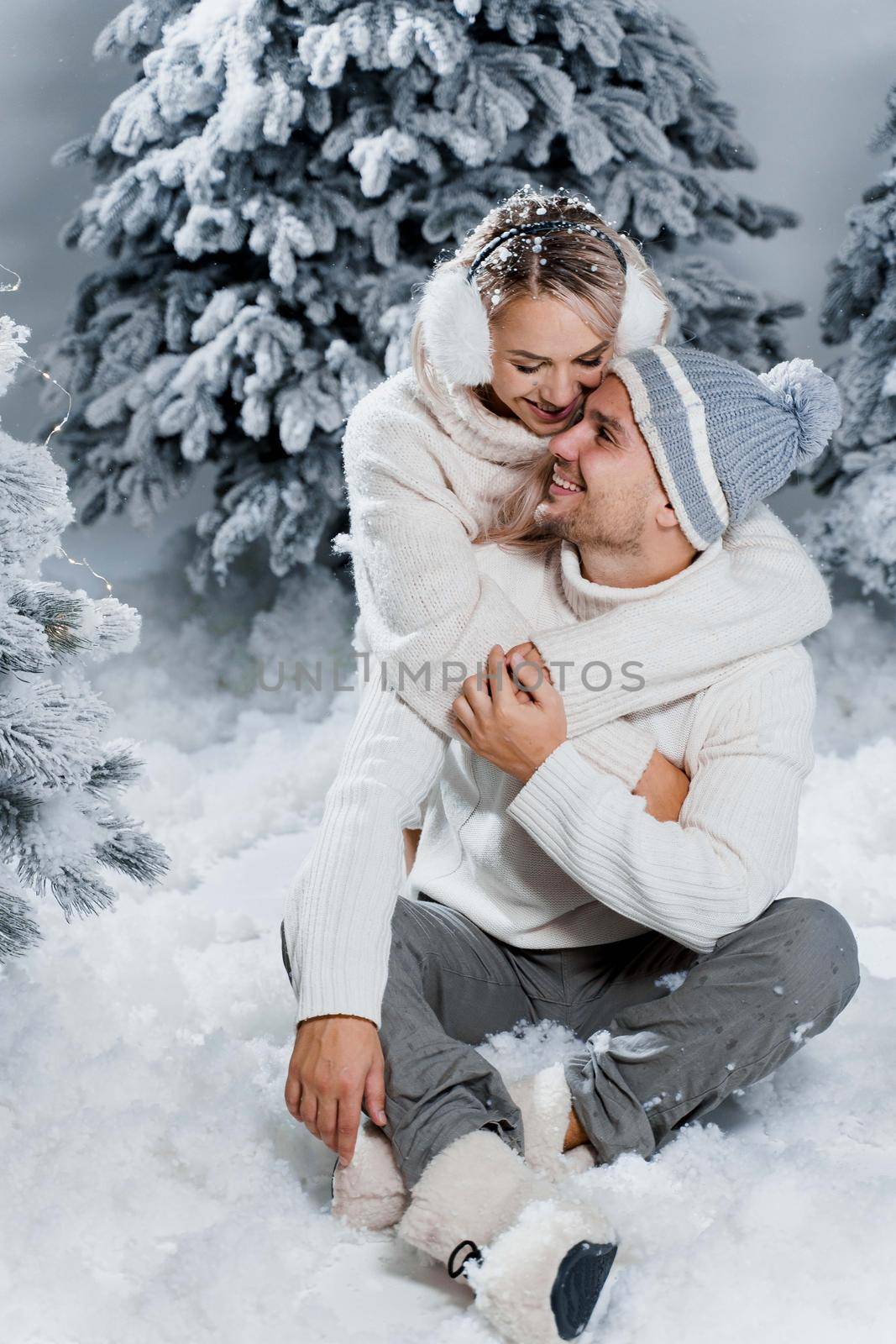 New year love story. Couple kisses and hugs. People weared wearing fur headphones, hats, white sweaters. Happy young couple hugs and kiss near christmas trees in winter day.