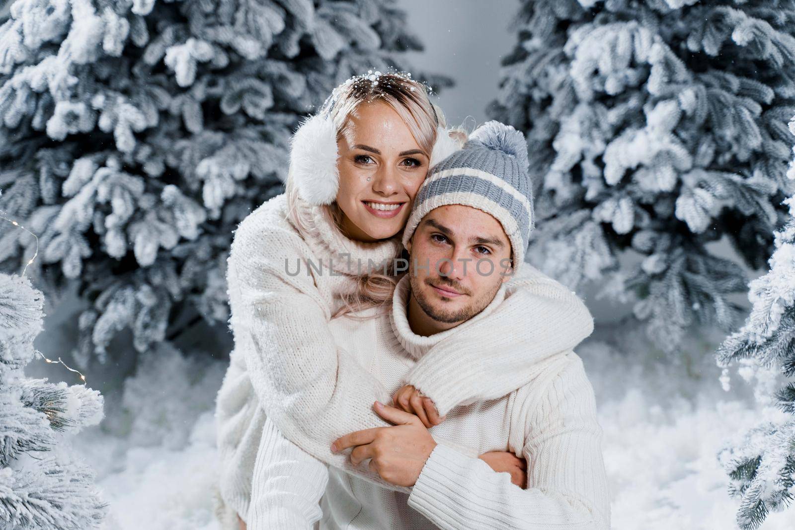 New year love story. Couple kisses and hugs. People weared wearing fur headphones, hats, white sweaters. Happy young couple hugs and kiss near christmas trees in winter day.