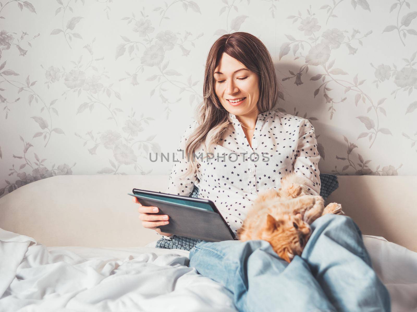 Woman sits on bed with tablet PC and cute ginger cat. She watches online TV series. Online video call or conference, distance learning, remote education. Self isolation during quarantine.