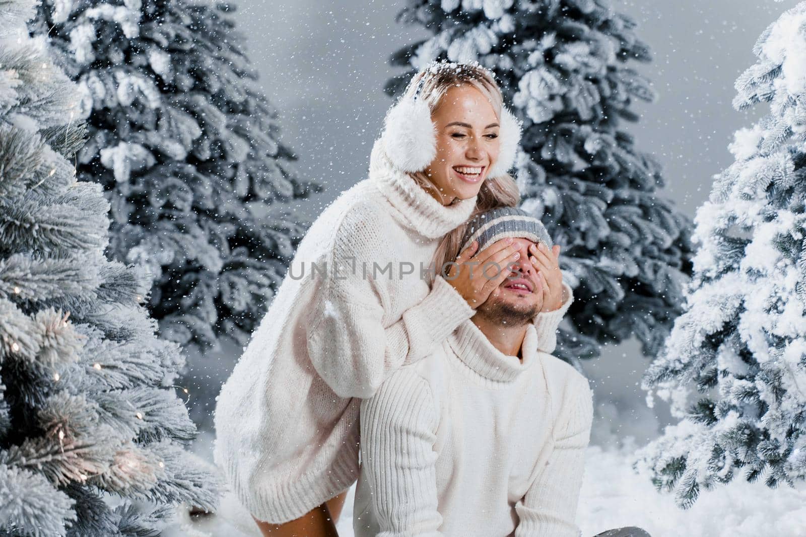 Winter love story with couple weared fur headphones, hats, white sweaters. Happy young couple hugs and kiss near christmas trees at the eve of new year celebration in winter day by Rabizo