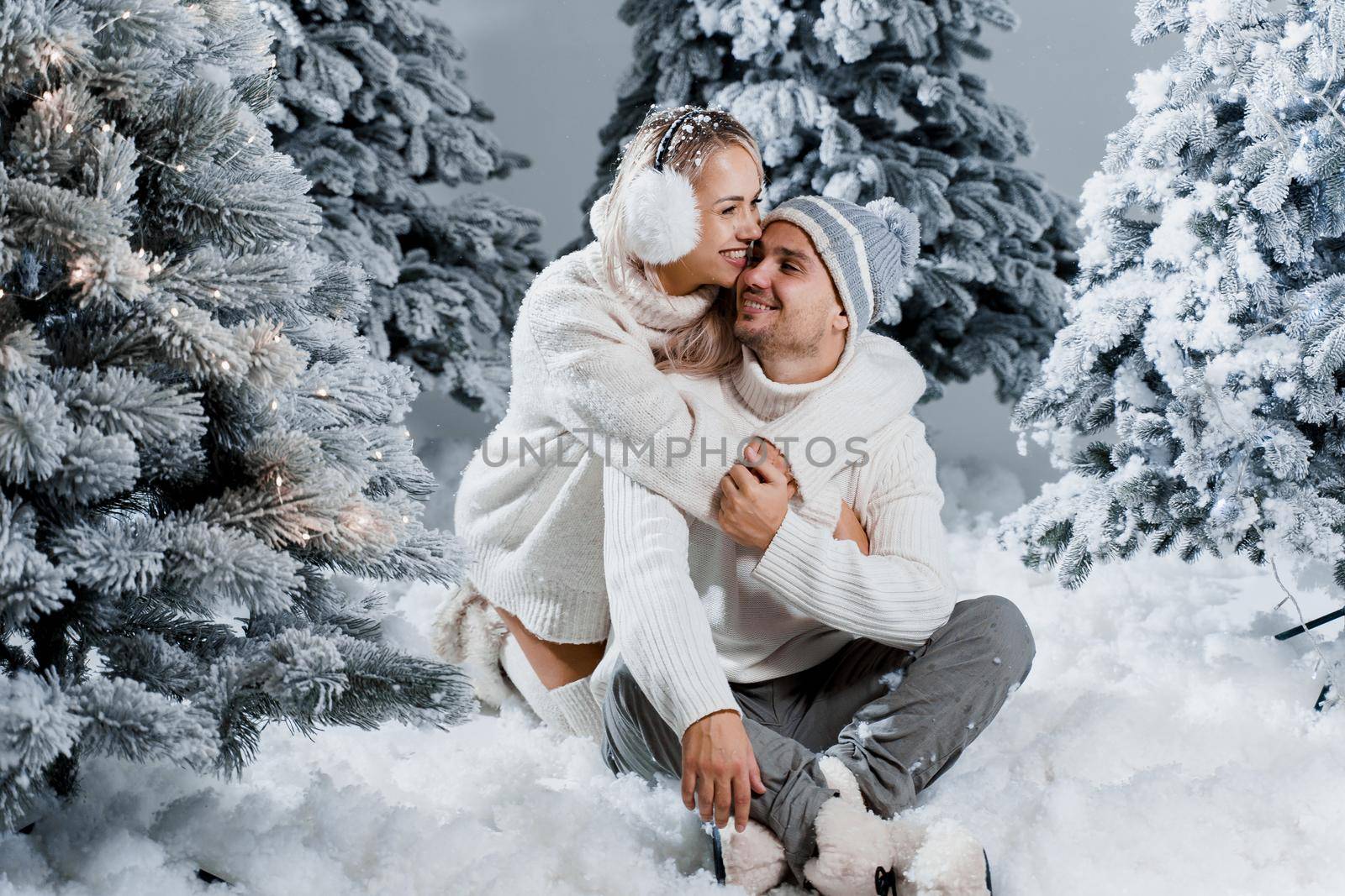 Couple kissing near christmass trees. Winter holidays. Love story of young couple weared white pullovers. Happy man and young woman hug each other by Rabizo