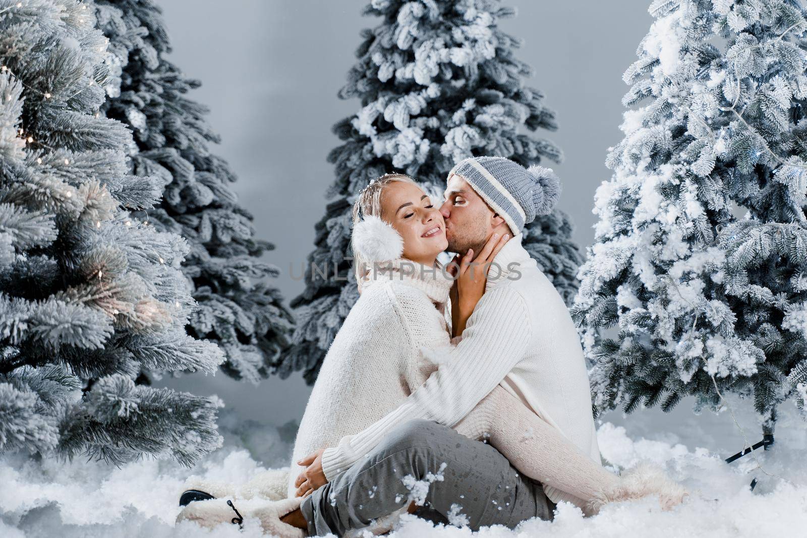 Young woman kissing her lovely man in cheek. Winter holidays. Love story of young couple weared white pullovers. Happy man and young woman hug each other.