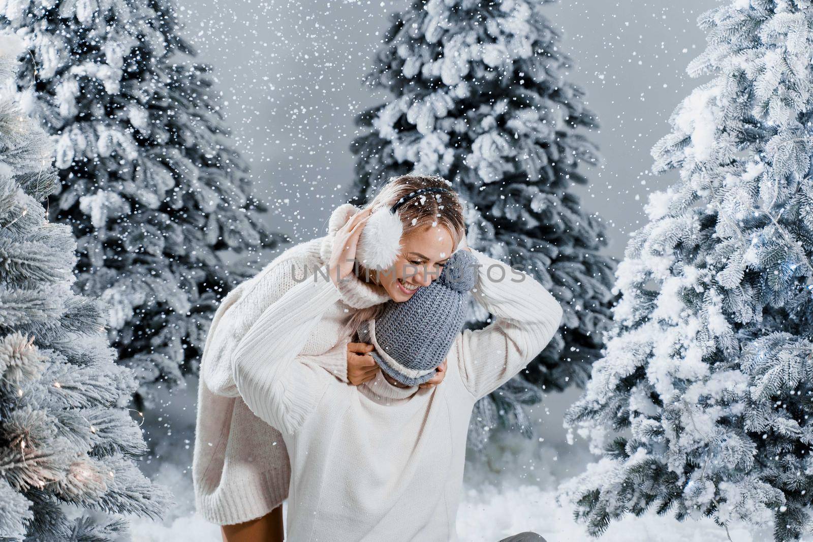New year love story. Couple kisses and hugs. People weared wearing fur headphones, hats, white sweaters. Happy young couple hugs and kiss near christmas trees in winter day.