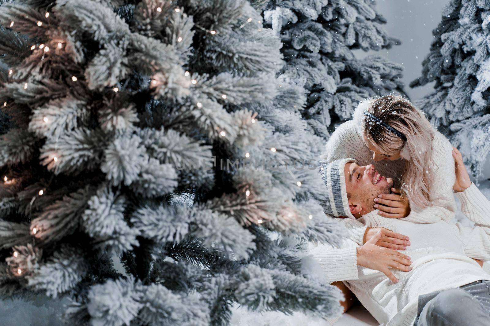 New year love story. Couple kisses and hugs. People weared wearing fur headphones, hats, white sweaters. Happy young couple hugs and kiss near christmas trees in winter day.