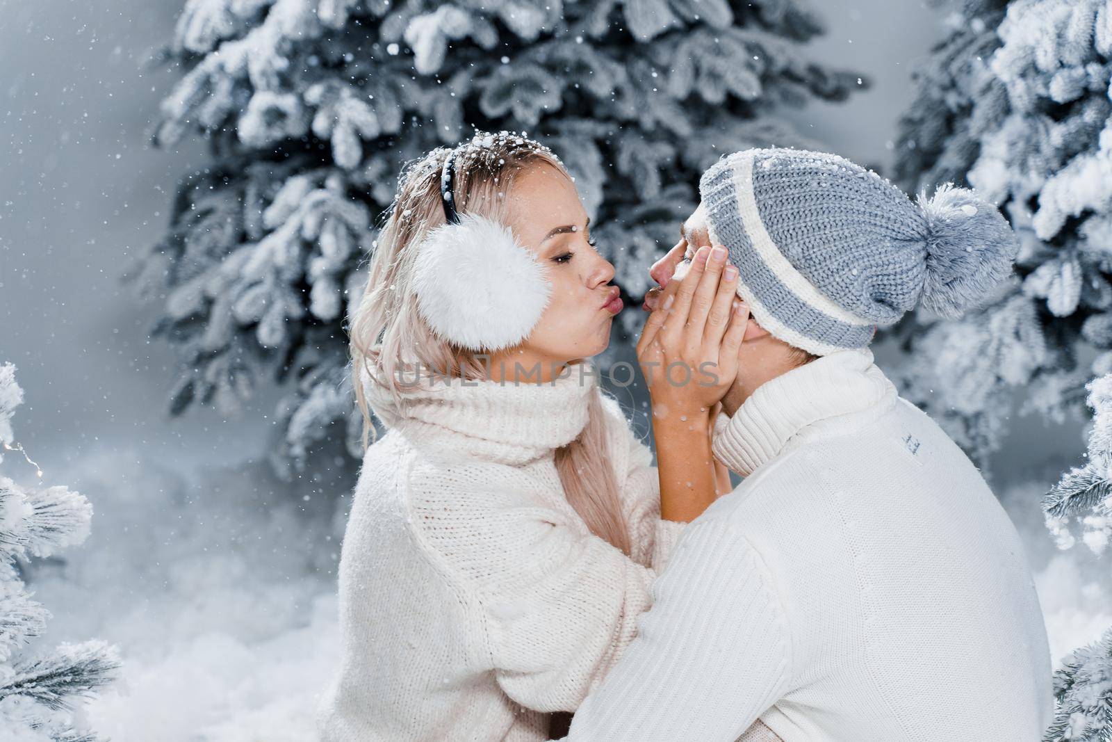 Couple couple laughing and having fun while snow falls near christmass trees. Winter holidays. Love story of young couple weared white pullovers. Happy man and young woman hug each other by Rabizo