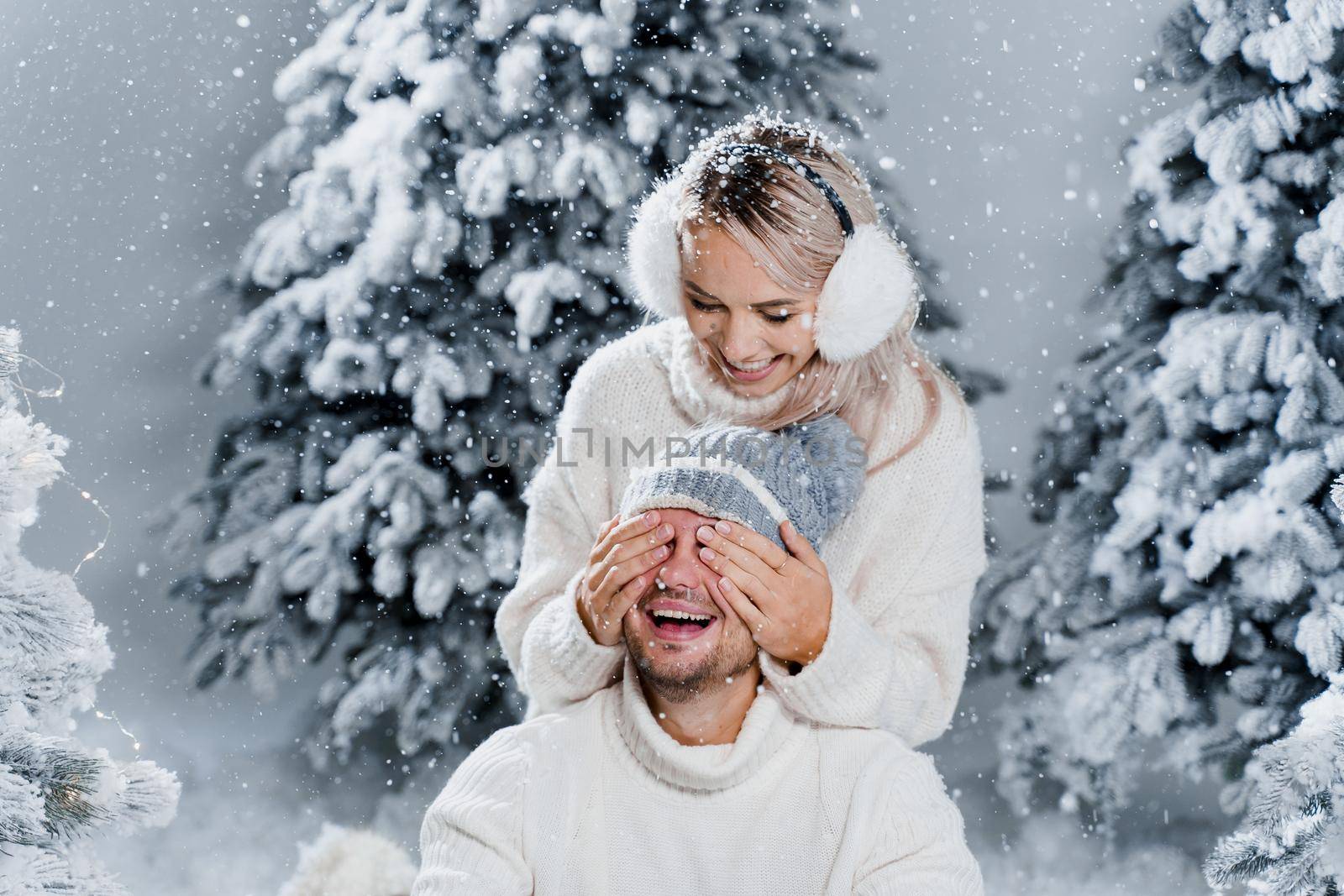 Couple couple laughing and having fun while snow falls near christmass trees. Winter holidays. Love story of young couple weared white pullovers. Happy man and young woman hug each other by Rabizo