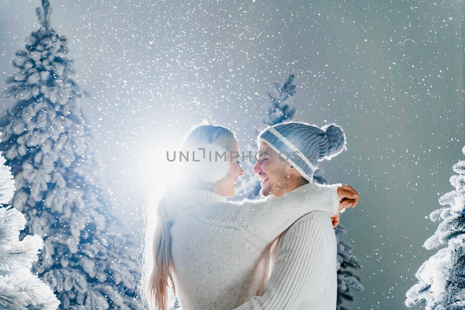 Happy young couple hugs and smiles and snow falls near christmas trees at the eve of new year celebration in winter day. Smiley man and woman weared white pullovers love each other. by Rabizo