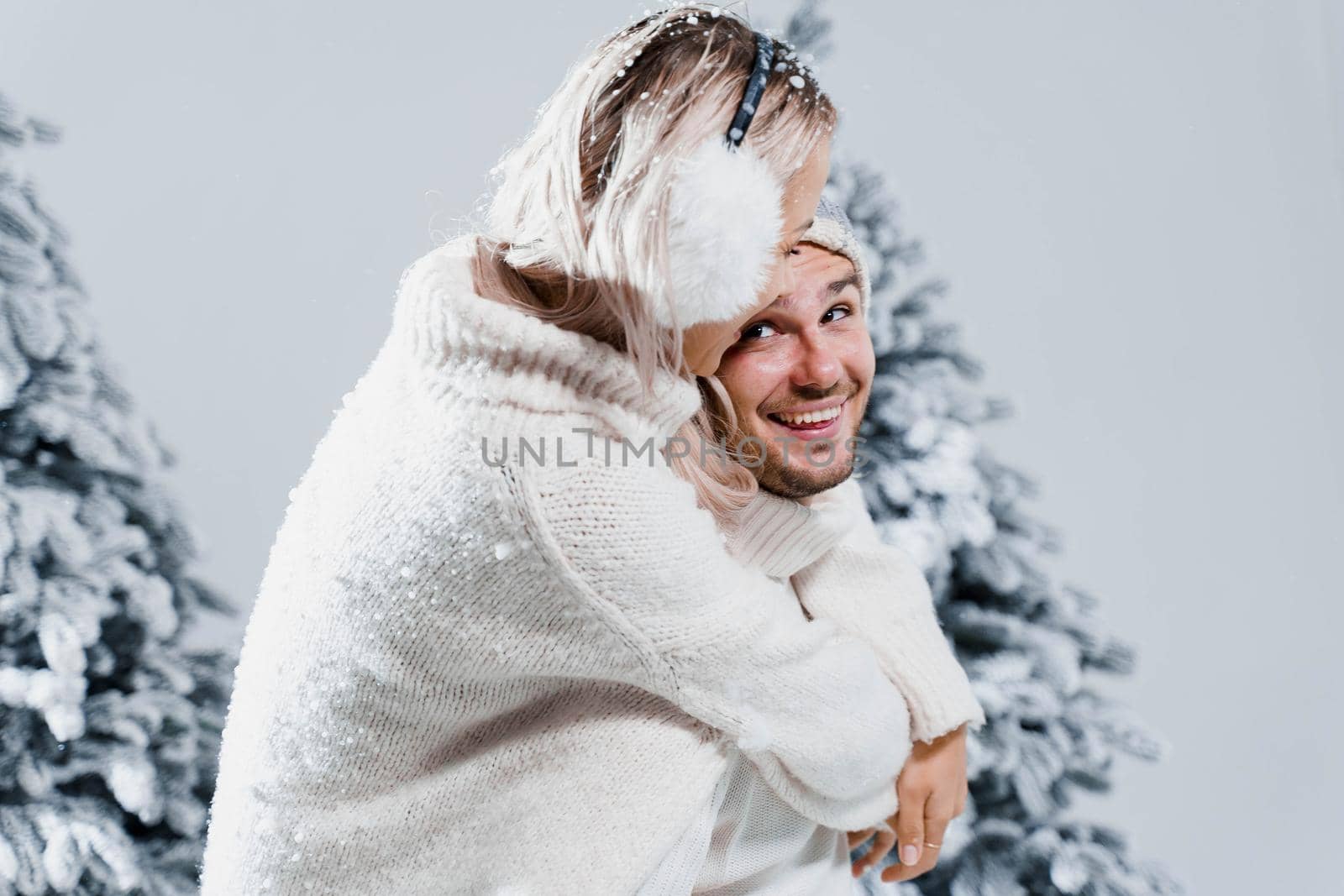 Winter love story with couple weared fur headphones, hats, white sweaters. Happy young couple hugs and kiss near christmas trees at the eve of new year celebration in winter day.