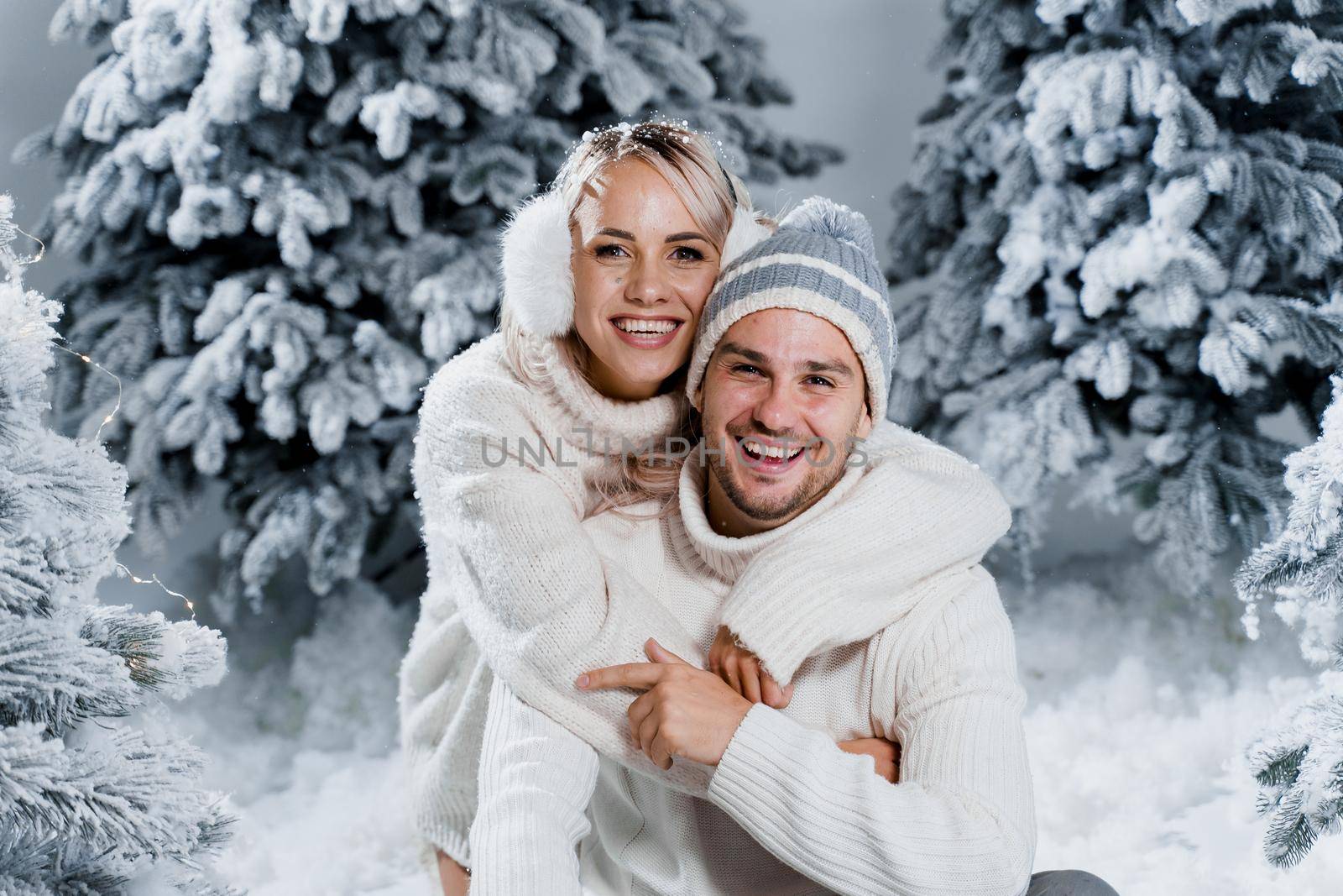 Couple couple laughing and having fun while snow falls near christmass trees. Winter holidays. Love story of young couple weared white pullovers. Happy man and young woman hug each other.