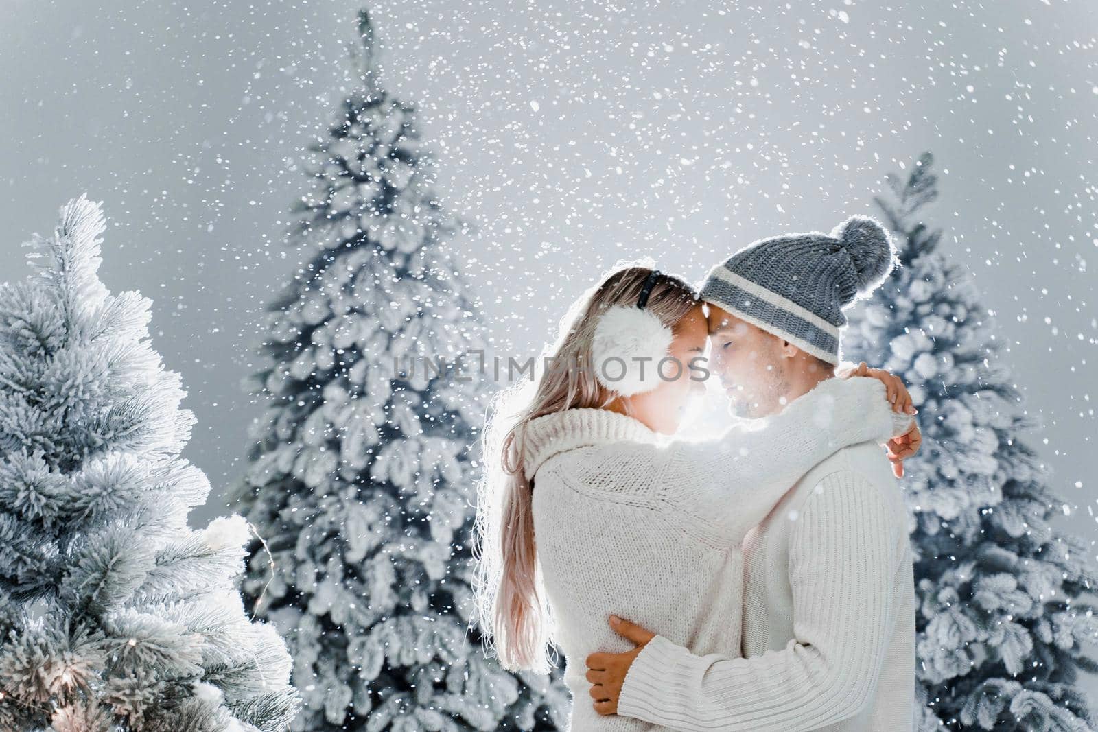 Happy young couple hugs and smiles and snow falls near christmas trees at the eve of new year celebration in winter day. Smiley man and woman weared white pullovers love each other