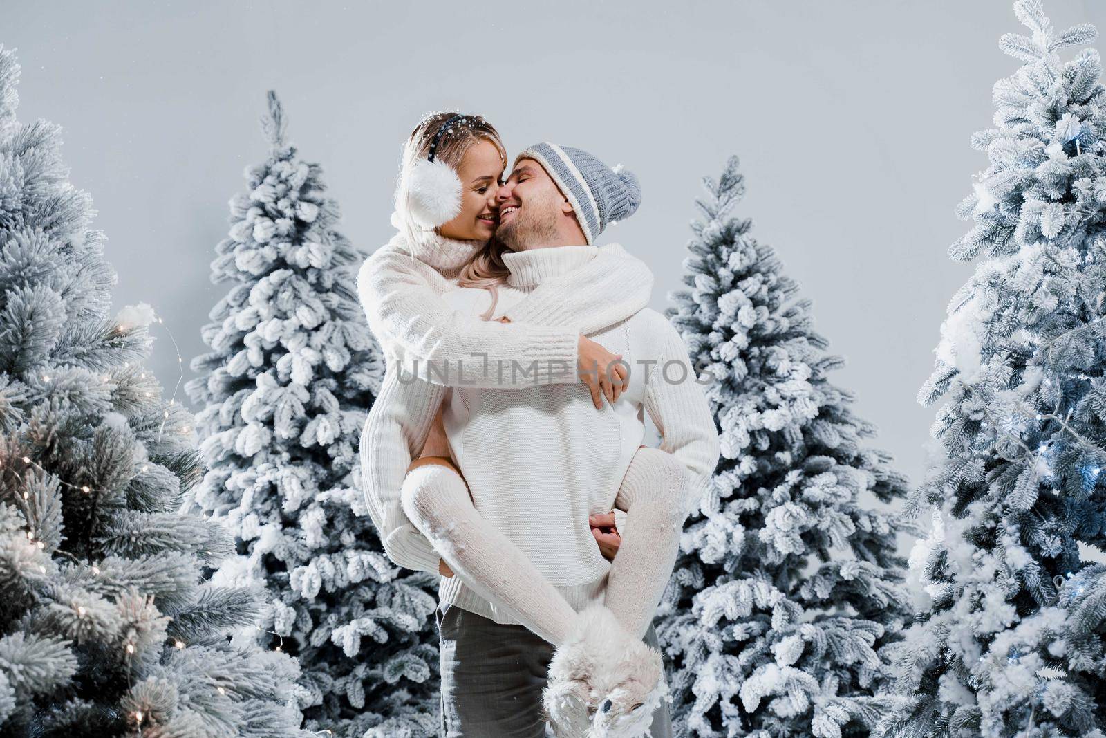 Couple kiss and hug. Man holds girl near christmas trees in winter day. New year celebration. People weared wearing fur headphones, hats, white sweaters.