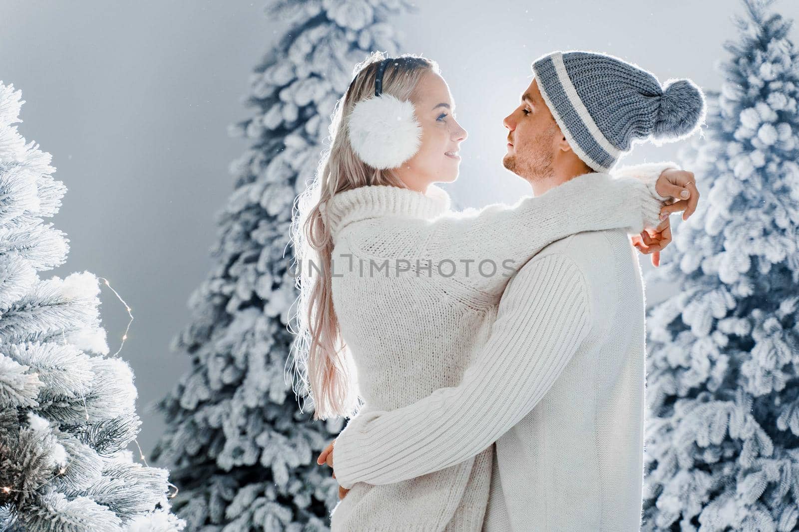 Winter love story with couple weared fur headphones, hats, white sweaters. Happy young couple hugs and kiss near christmas trees at the eve of new year celebration in winter day.