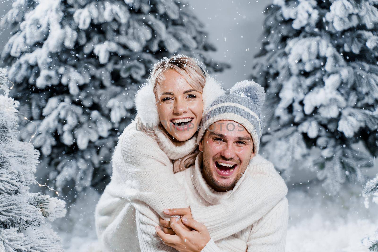 Couple couple laughing and having fun while snow falls near christmass trees. Winter holidays. Love story of young couple weared white pullovers. Happy man and young woman hug each other.