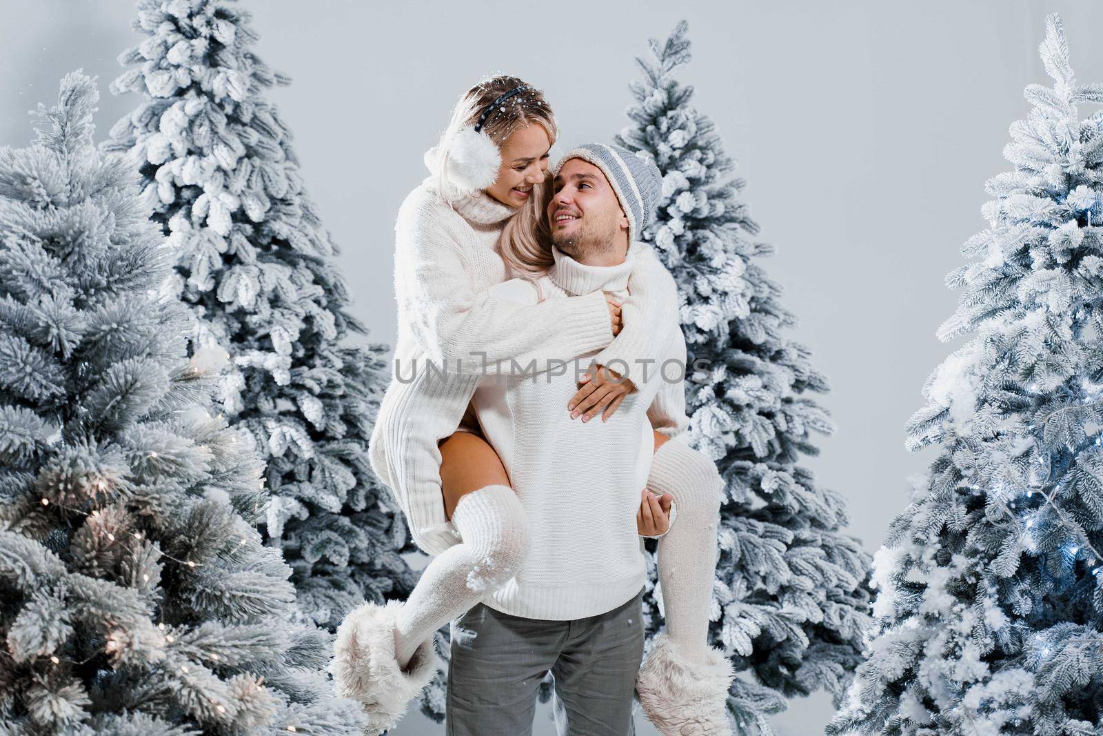 Couple kiss and hug. Man holds girl near christmas trees in winter day. New year celebration. People weared wearing fur headphones, hats, white sweaters by Rabizo