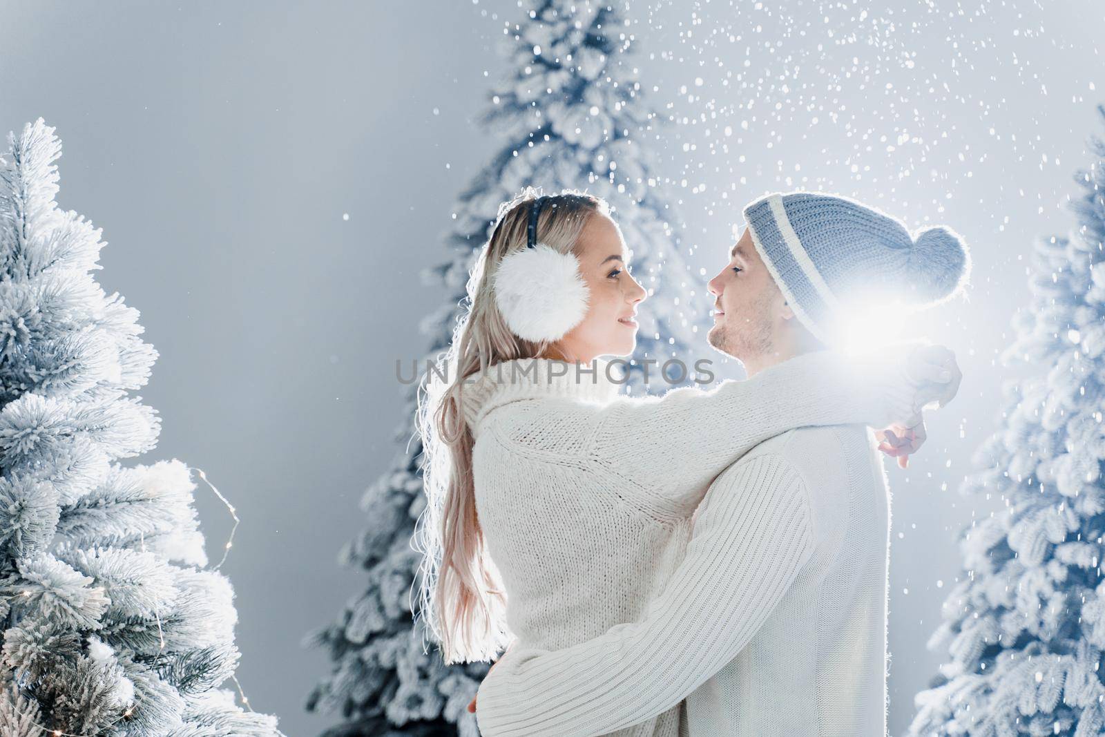 Couple kissing while snow falls near christmass trees. Winter holidays. Love story of young couple weared white pullovers. Happy man and young woman hug each other.